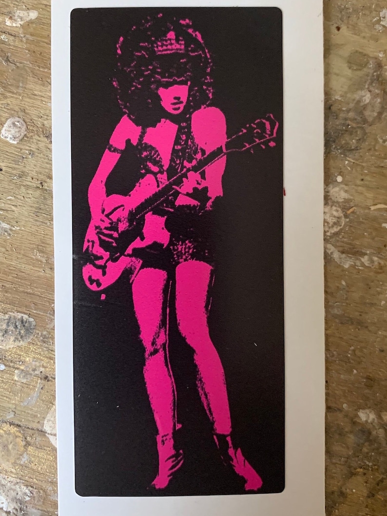 Op de afbeelding: Een zwart-wit afbeelding van een persoon die gitaar speelt. De persoon draagt een roze outfit en heeft een grote afro. De gitaar is lichtbruin.
