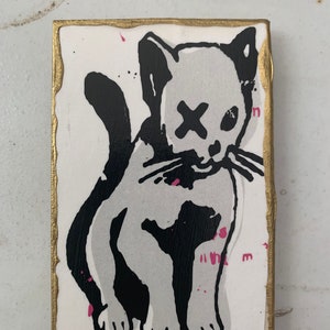 May include: A black and white illustration of a cat with an X over its eye. The cat is painted on a white wooden block with a gold border. There are pink paint splatters on the background.