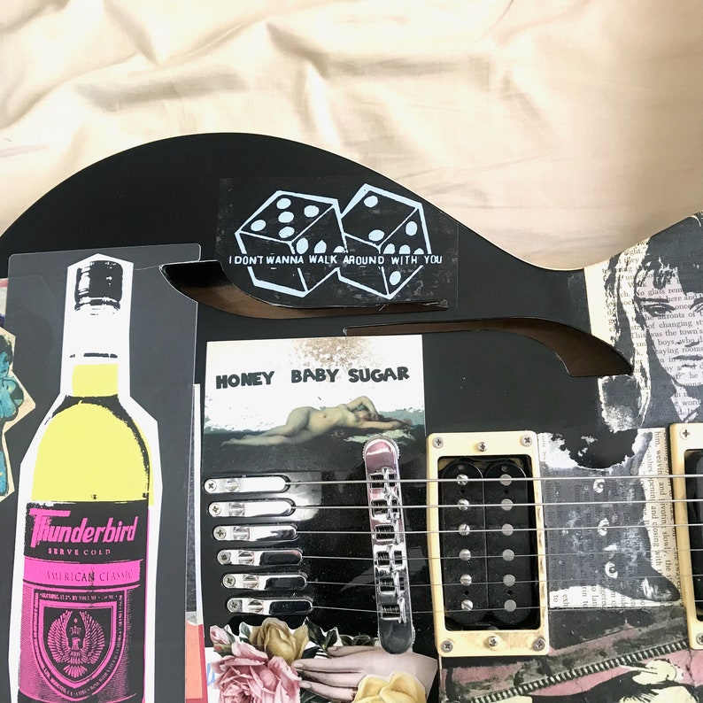 May include: A black electric guitar with a white sticker that says "I don't wanna walk around with you" and two dice. There is a sticker on the guitar that says "Honey Baby Sugar" with a woman lying on a beach. There is a bottle of Thunderbird American Classic liquor in the image.