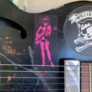 Op de afbeelding: Een zwarte elektrische gitaar met een sticker van een vrouw in roze die gitaar speelt. De sticker zegt "I Will Cut You".