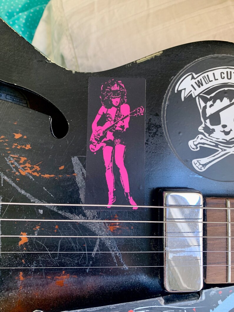 Op de afbeelding: Een zwarte elektrische gitaar met een sticker van een vrouw in roze die gitaar speelt. De sticker zegt "I Will Cut You".