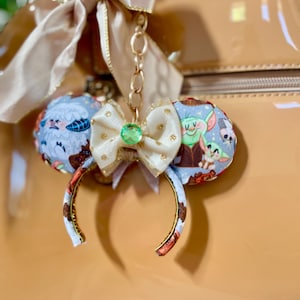 May include: A miniature mouse ear headband keychain featuring a light blue fabric with a cartoon character print. The headband is adorned with a small cream-colored bow with gold glitter accents and a central green gem, attached to a gold chain and ribbon.