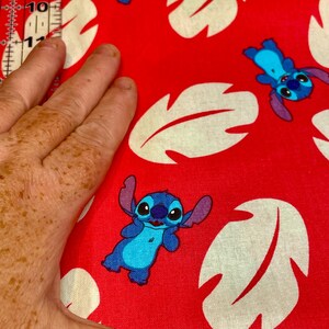 May include: Red fabric with a repeating pattern of blue cartoon characters and cream-coloured leaf shapes. The fabric is being measured with a ruler. The cartoon character has large ears and a friendly expression.