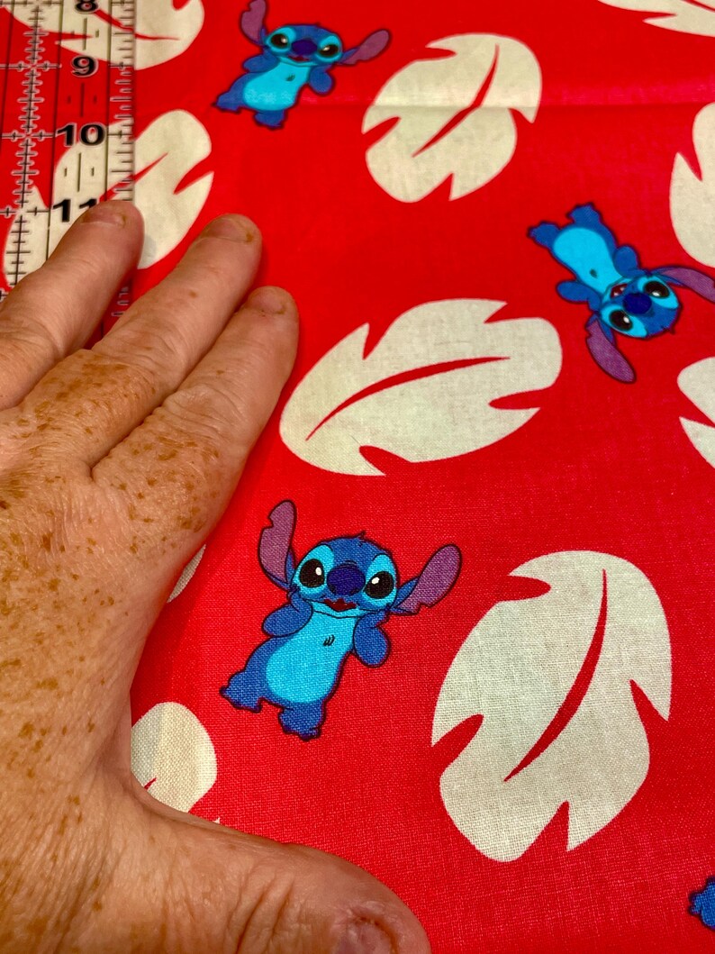 May include: Red fabric with a repeating pattern of blue cartoon characters and cream-coloured leaf shapes. The fabric is being measured with a ruler. The cartoon character has large ears and a friendly expression.