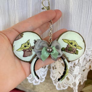 May include: A pair of light green mouse ears with a silver and green bow and a silver chain attached. The ears are decorated with a pattern of a green alien character.
