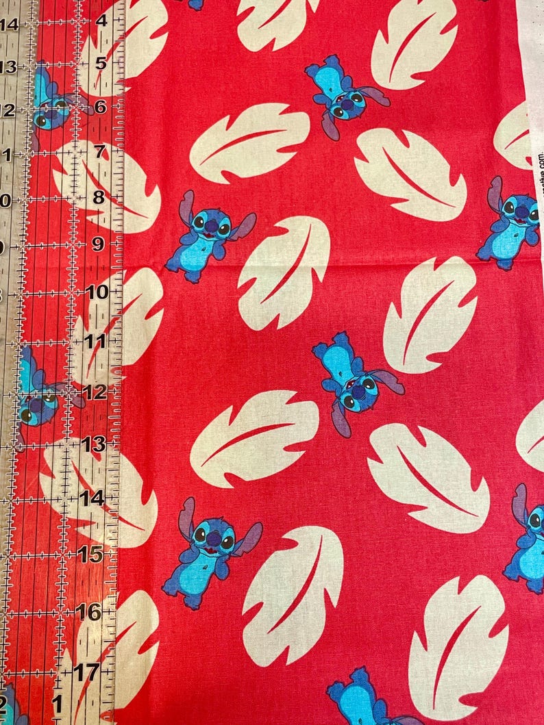 May include: Red fabric with a repeating pattern of blue and white Stitch characters and large cream-colored leaf shapes. A ruler with inch measurements is visible along the left side of the fabric.