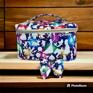 Könnte beinhalten: Eine weiße, ovale Kosmetiktasche mit Reißverschluss und goldenem Griff. Die Tasche ist mit einem bunten Druck von Disney-Charakteren in Silhouette bedeckt. Die Tasche liegt auf einer Holzoberfläche mit einer passenden Schleife.