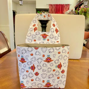 May include: White tote bag with a repeating pattern of cartoon characters in orange, red, and blue. The bag has a matching handle and a beige interior. The bag is sitting on a wooden table.