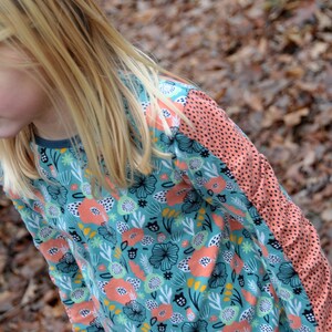 Puede incluir: Una ni&ntilde;a lleva una camisa de manga larga con un estampado floral azul verdoso, rosa y blanco. La camisa tiene un panel de color melocot&oacute;n en la manga derecha con lunares negros.