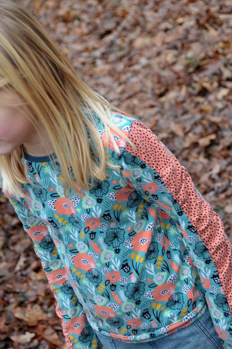 Puede incluir: Una ni&ntilde;a lleva una camisa de manga larga con un estampado floral azul verdoso, rosa y blanco. La camisa tiene un panel de color melocot&oacute;n en la manga derecha con lunares negros.