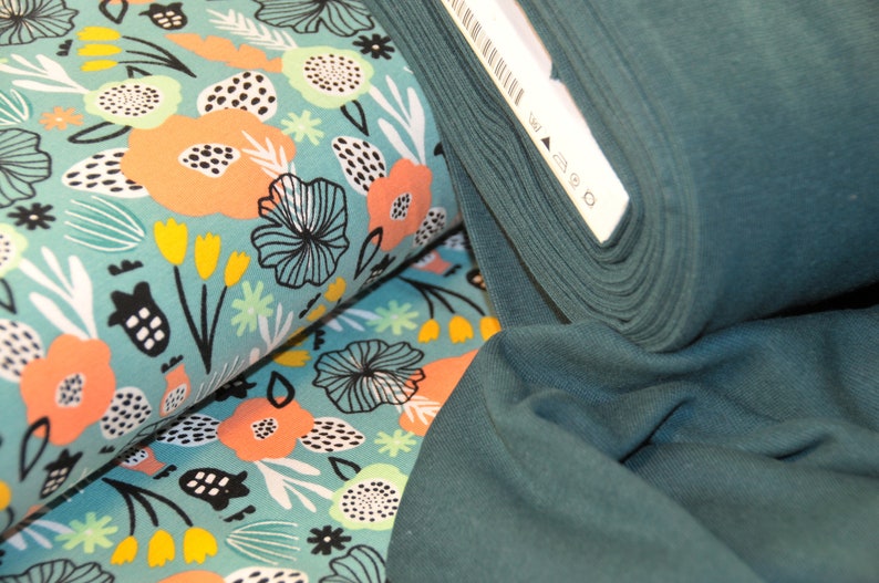 Puede incluir: Un rollo de tela de color verde azulado con estampado floral que presenta flores rosas, amarillas y blancas sobre un fondo azul claro.