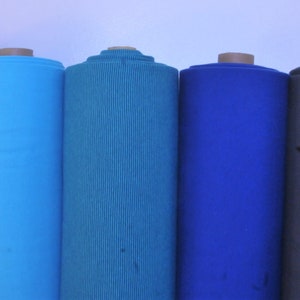 Könnte beinhalten: Fünf Stoffrollen in verschiedenen Blautönen und Grau. Die Stoffrollen sind horizontal von links nach rechts angeordnet. Die Stoffrollen sind alle gleich groß und in braunes Papier eingewickelt.