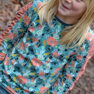 Puede incluir: Una camisa de manga larga con fondo azul verdoso y un estampado floral colorido. Las mangas tienen un ribete de color melocot&oacute;n con lunares blancos.