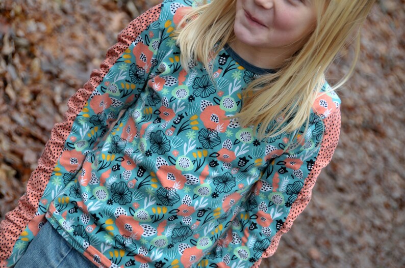 Puede incluir: Una camisa de manga larga con fondo azul verdoso y un estampado floral colorido. Las mangas tienen un ribete de color melocot&oacute;n con lunares blancos.