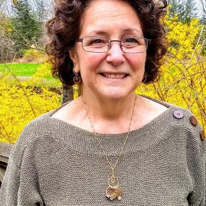 May include: A woman with curly brown hair is wearing a gray sweater with a gold necklace and a pendant. She is smiling and looking at the camera. The background is a blurry image of trees and yellow flowers.
