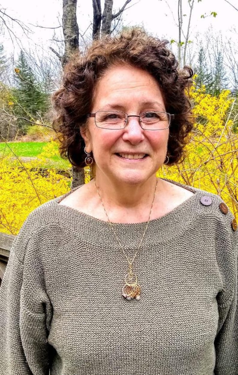 May include: A woman with curly brown hair is wearing a gray sweater with a gold necklace and a pendant. She is smiling and looking at the camera. The background is a blurry image of trees and yellow flowers.