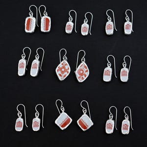 May include: A collection of silver earrings with various ceramic designs. The earrings feature rectangular, oval, and diamond shapes with red and white patterns. The earrings are displayed against a black background.