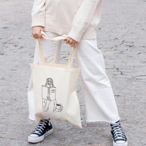 May include: A white canvas tote bag with a black line drawing of a person reading a book and holding a cup of coffee. The text on the book says "Read" with a squiggly line underneath.