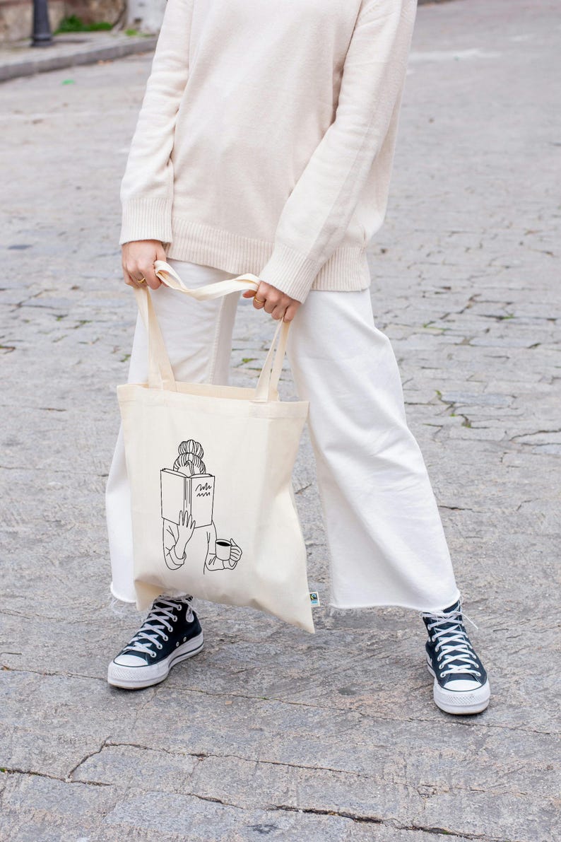 May include: A white canvas tote bag with a black line drawing of a person reading a book and holding a cup of coffee. The text on the book says "Read" with a squiggly line underneath.