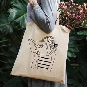 May include: A white tote bag with a black line drawing of a person reading a book. The book has the word "Mmm" written on the cover.