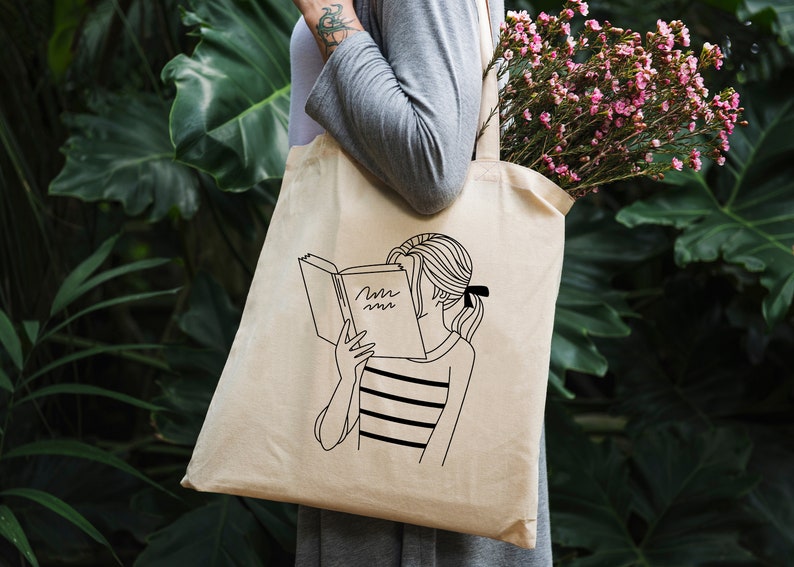 May include: A white tote bag with a black line drawing of a person reading a book. The book has the word "Mmm" written on the cover.