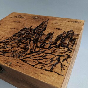 May include: A wooden box with a hinged lid and a metal latch. The top of the box is decorated with a woodburned image of Hogwarts castle.