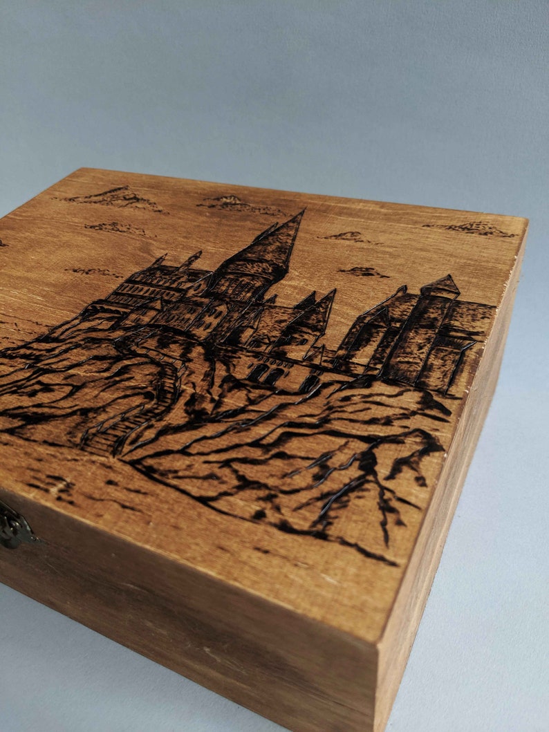 May include: A wooden box with a hinged lid and a metal latch. The top of the box is decorated with a woodburned image of Hogwarts castle.