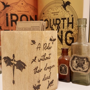 May include: A wooden bookend with a dragon design and the text "A Rider without their dragon is dead". The bookend is sitting on a table with two glass bottles and two books in the background.