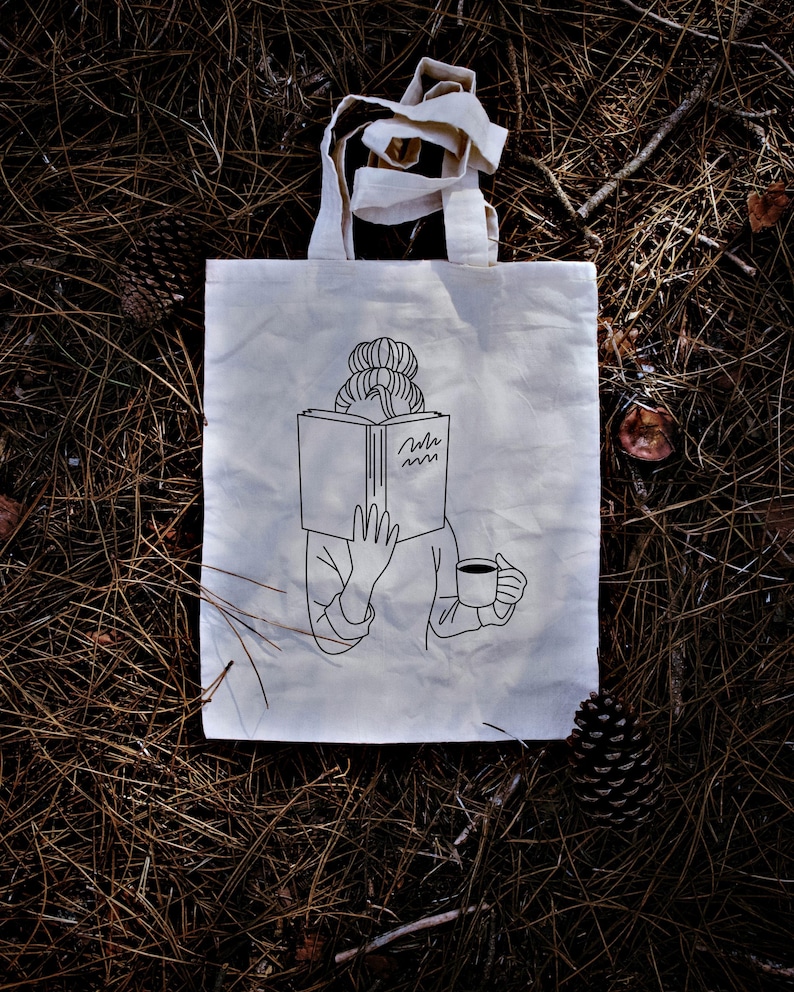 May include: A white tote bag with a black line drawing of a person reading a book and holding a cup of coffee. The person has a bun in their hair and the book has the word "mmm" written on it.