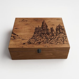 May include: A wooden box with a hinged lid and a metal latch. The top of the box features a woodburned image of Hogwarts castle from the Harry Potter series.