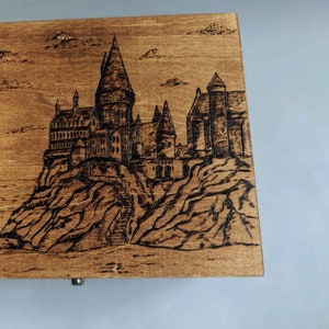 May include: A wooden box with a burnt-in image of Hogwarts castle, a fictional school of witchcraft and wizardry from the Harry Potter series.