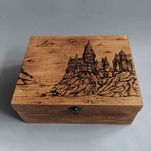 May include: A wooden box with a hinged lid and a metal latch. The top of the box is decorated with a woodburned image of Hogwarts castle.