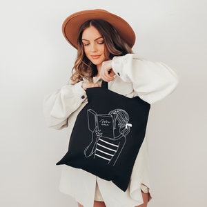 May include: A black tote bag with a white line drawing of a person reading a book. The book says "Lost in a book".