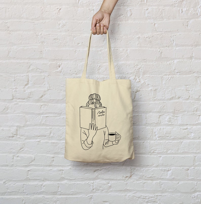 May include: A beige tote bag with a black line drawing of a person reading a book and holding a cup of coffee. The text on the book reads "Read more".