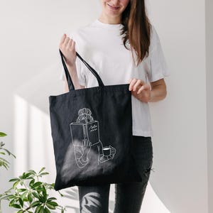 May include: A black tote bag with a white outline of a person reading a book and holding a cup. The text on the book says "The book".