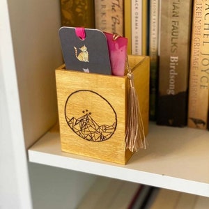 May include: A wooden bookend with a geometric mountain design and five stars. The bookend has a tassel and a small compartment for holding bookmarks. The compartment contains a black bookmark with a gold cat illustration and a pink bookmark.
