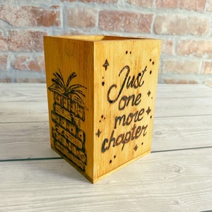 May include: A wooden pencil holder with a stack of books and the text "Just one more chapter" burned into the wood.