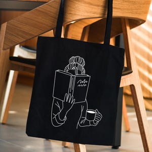 May include: Black tote bag with a white illustration of a person reading a book and holding a cup of coffee. The text on the book reads "Hello".