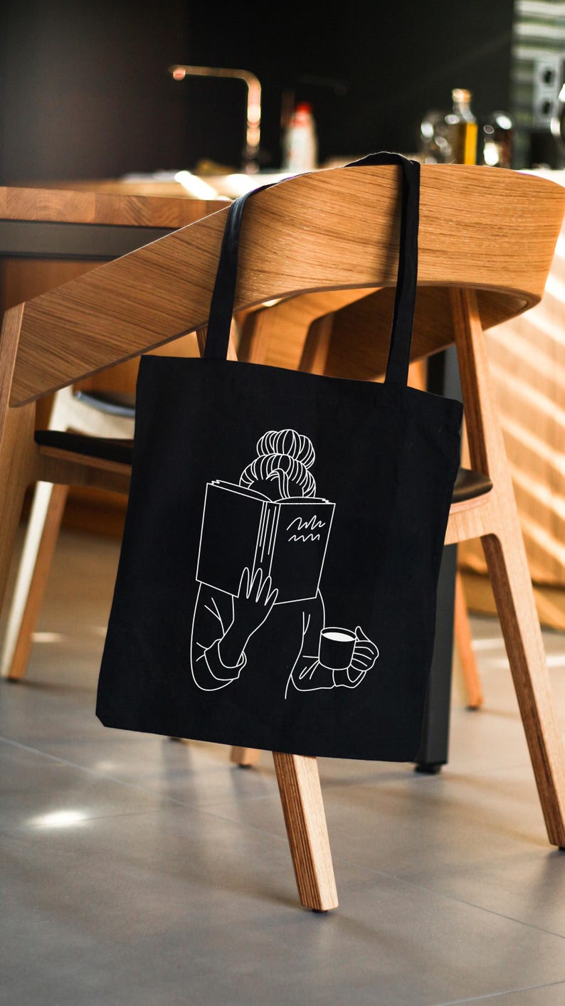 May include: Black tote bag with a white illustration of a person reading a book and holding a cup of coffee. The text on the book reads "Hello".