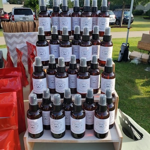 May include: A display of brown glass spray bottles with white labels featuring a cartoon mustache and the text "The Mustache Club" on a wooden tiered stand. The bottles are arranged in rows on the stand.