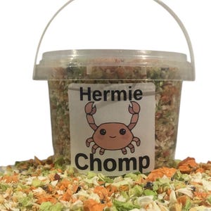 May include: A clear plastic bucket filled with a colorful mix of dried vegetables and herbs. The bucket has a white label with the text "Hermie Chomp" and a cartoon crab illustration. The mix is also scattered around the base of the bucket.