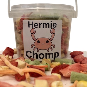 May include: A clear plastic bucket filled with a variety of dried fruits and vegetables. The bucket has a label that says "Hermie Chomp" and features a cartoon crab.