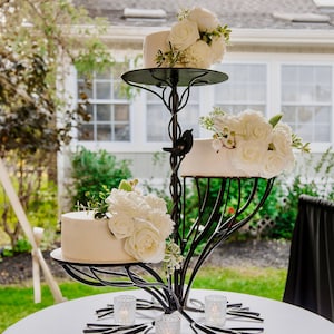 May include: A three-tiered wedding cake stand made of black metal with a tree branch design. The stand is decorated with white flowers and holds three white cakes. The stand is on a white tablecloth with four small glass candle holders.