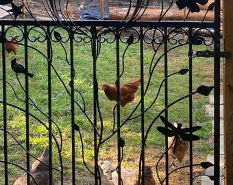 Op de afbeelding: Een zwart metalen tuinhek met decoratieve silhouetten van vlinders, vogels en een libel geïntegreerd in de verticale spijlen. Het hek heeft een boogvormige bovenkant en gebogen metalen ranken met bladaccenten door het frame.