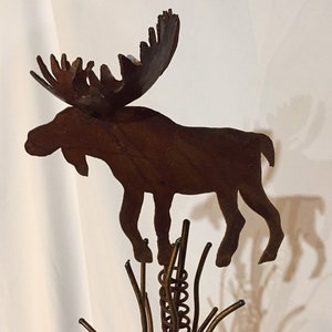 May include: A metal moose silhouette with large antlers, standing on a coiled spring and metal branches. The moose is a dark brown color and has a rustic, weathered look.