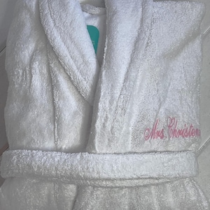 May include: White bathrobe with a shawl collar and a pink embroidered heart and the text "Mrs. Christensen".