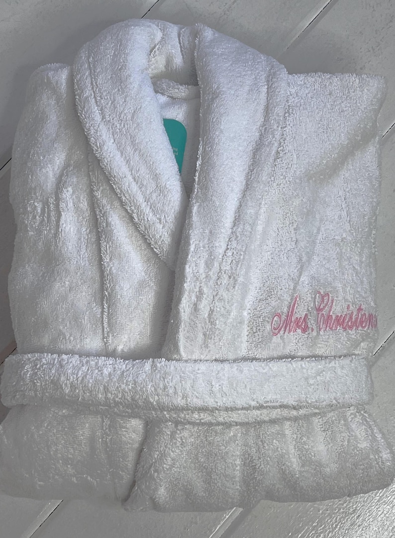 May include: White bathrobe with a shawl collar and a pink embroidered heart and the text "Mrs. Christensen".