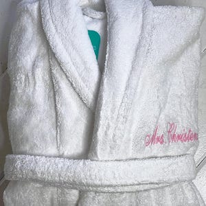 May include: White bathrobe with a shawl collar and a pink embroidered name "Mrs. Christon" on the right side.