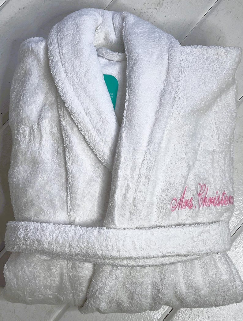 May include: White bathrobe with a shawl collar and a pink embroidered name "Mrs. Christon" on the right side.