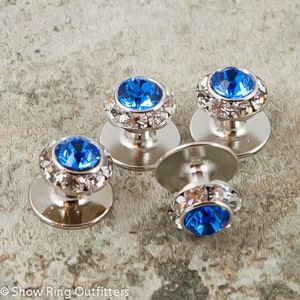 May include: Four silver-toned shirt studs with a bright blue center stone surrounded by clear crystals. The studs have a round, flat base and a domed top. The gemstones are set in a silver-colored metal.